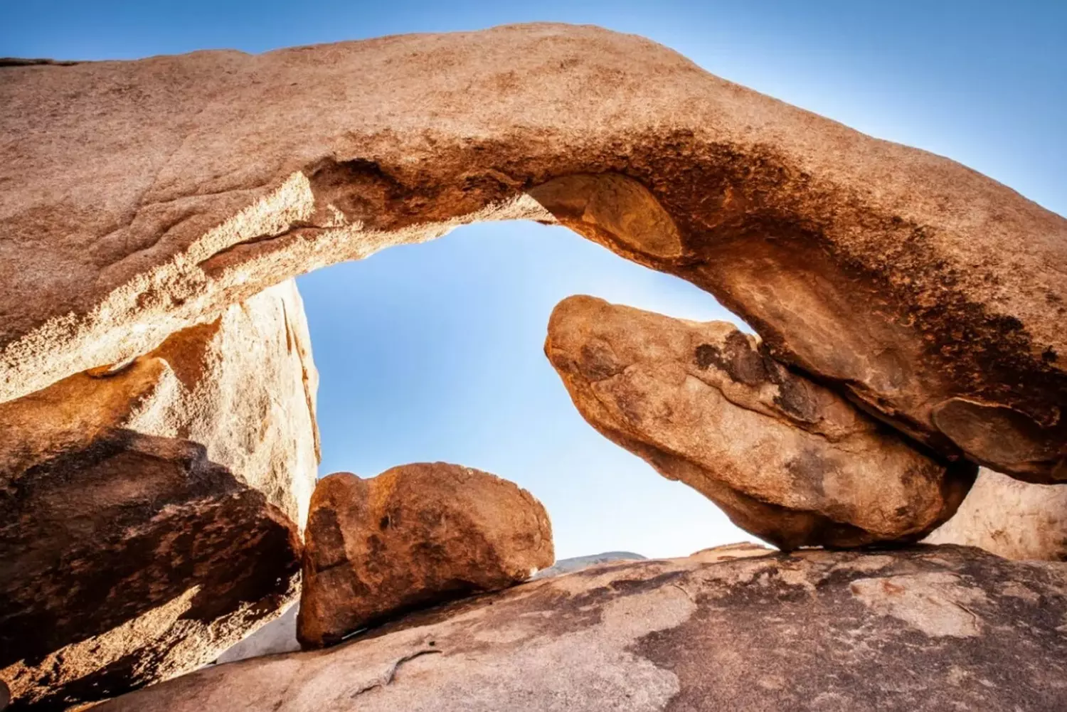 Nature's Sculpture: Arch Rock in Joshua Tree National Park A stunning natural granite arch formation at Joshua Tree National Park, perfectly framing the desert landscape and blue sky, with hikers visible for scale showing its massive size