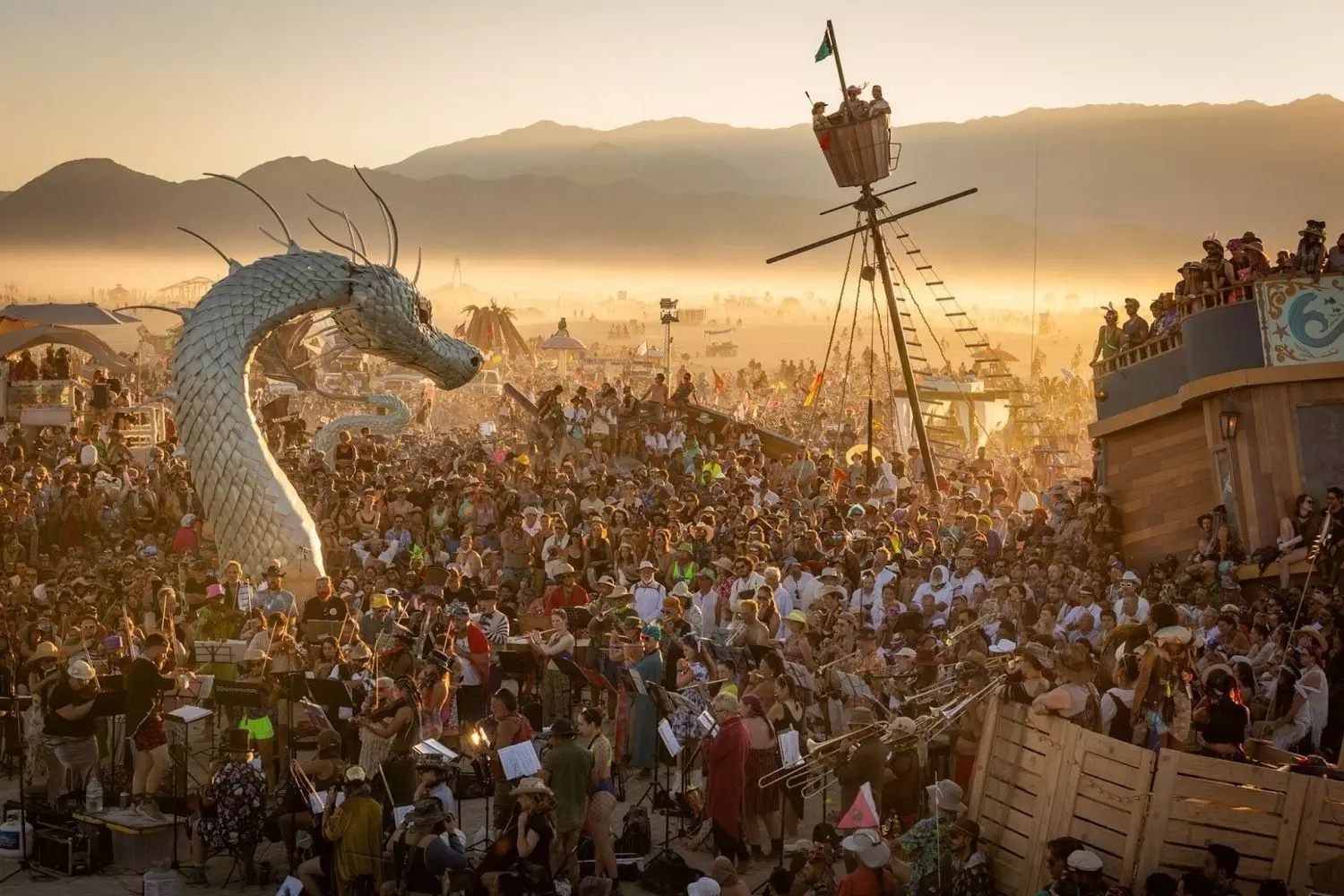 Дракон на Burning Man: Толпа, огонь и магия пустыни Огромная толпа на фестивале Burning Man вокруг гигантской арт-инсталляции дракона в пустыне, в воздухе — пыль и атмосфера праздника
