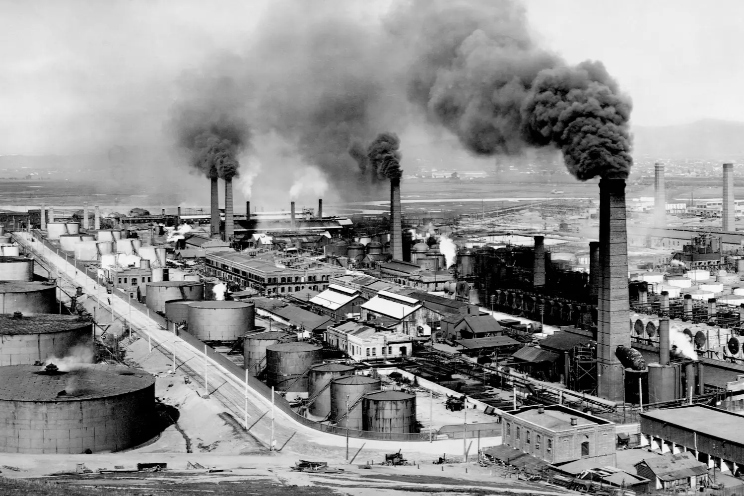 Historic Oil Refinery in the USA: Industrial Legacy of the 20th Century A black-and-white photo of an early 20th-century American oil refinery with smokestacks, pipelines, and storage tanks, showcasing the dawn of the petroleum industry