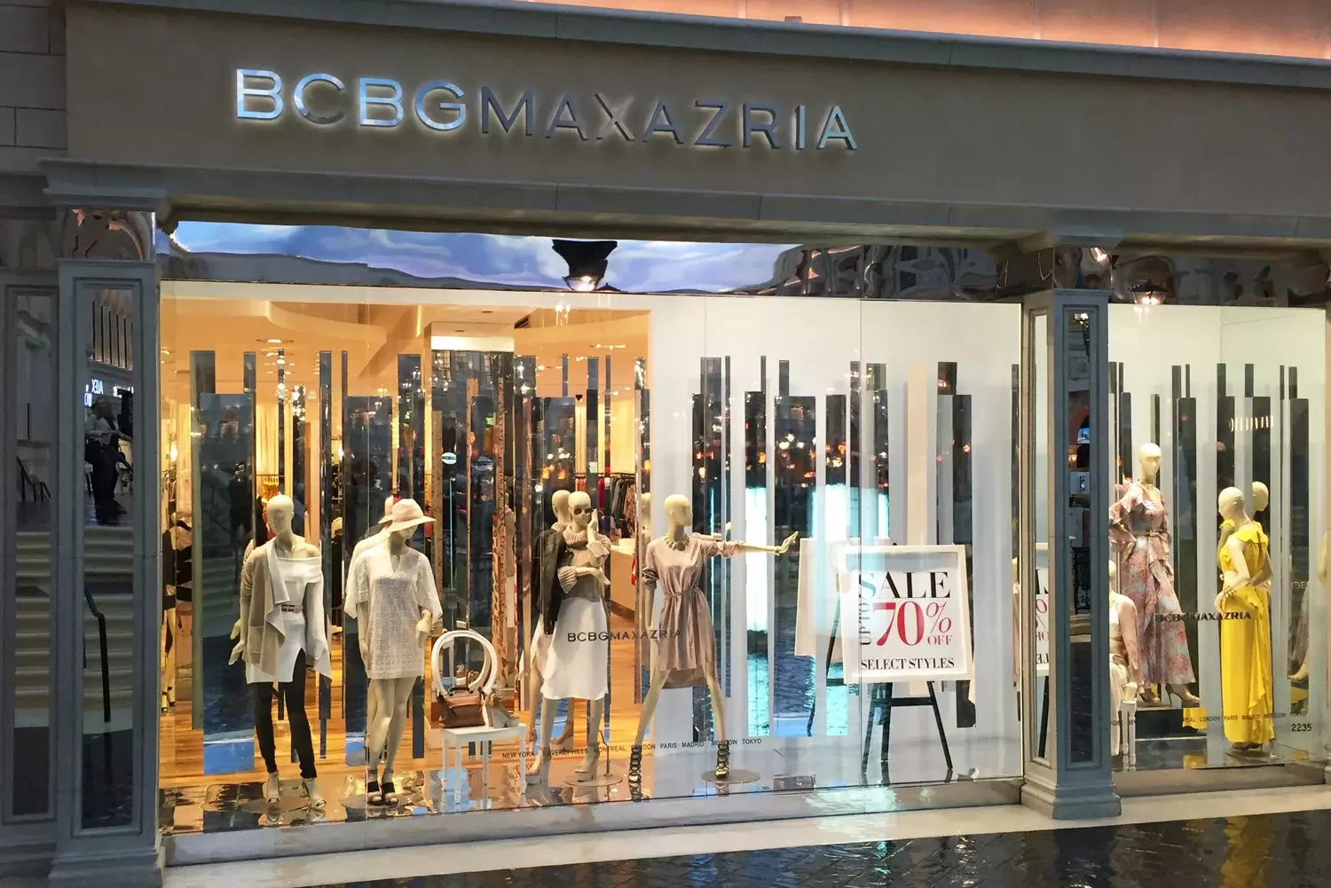 Магазин BCBG: Французский шик с калифорнийским настроением Фото бутика BCBG с изысканными витринами, демонстрирующими платья в пол, стильные ансамбли и фирменные женственные детали