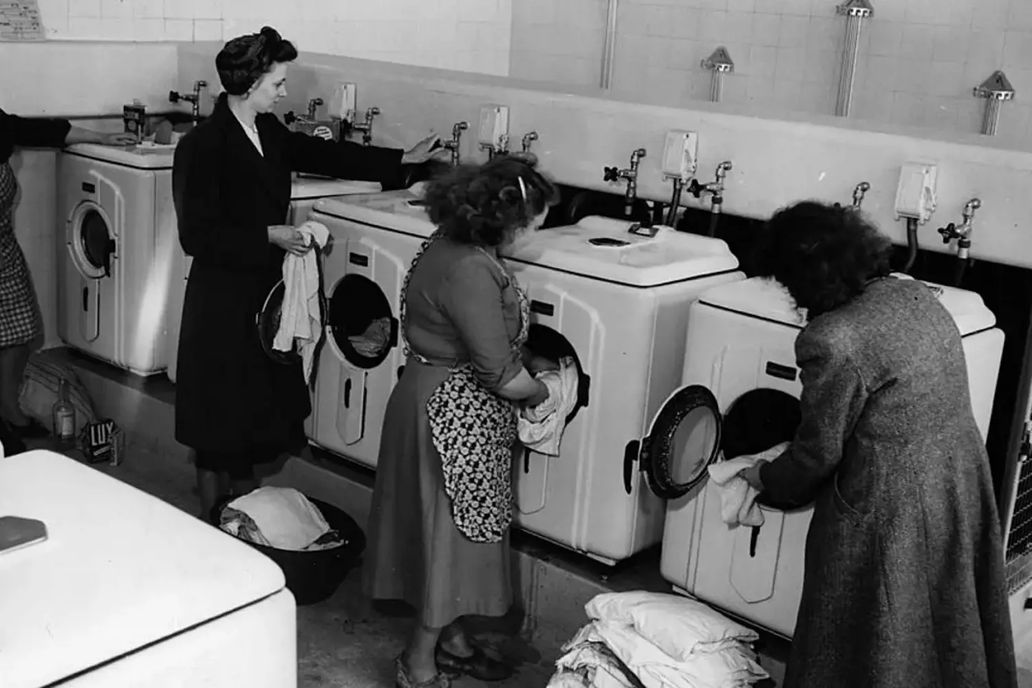Vintage Laundromat in the USA: A Glimpse into 20th Century Everyday Life Black-and-white photo of a mid-century American laundromat with retro washing machines, coin-operated dryers, and customers folding clothes — a snapshot of 1950s domestic culture