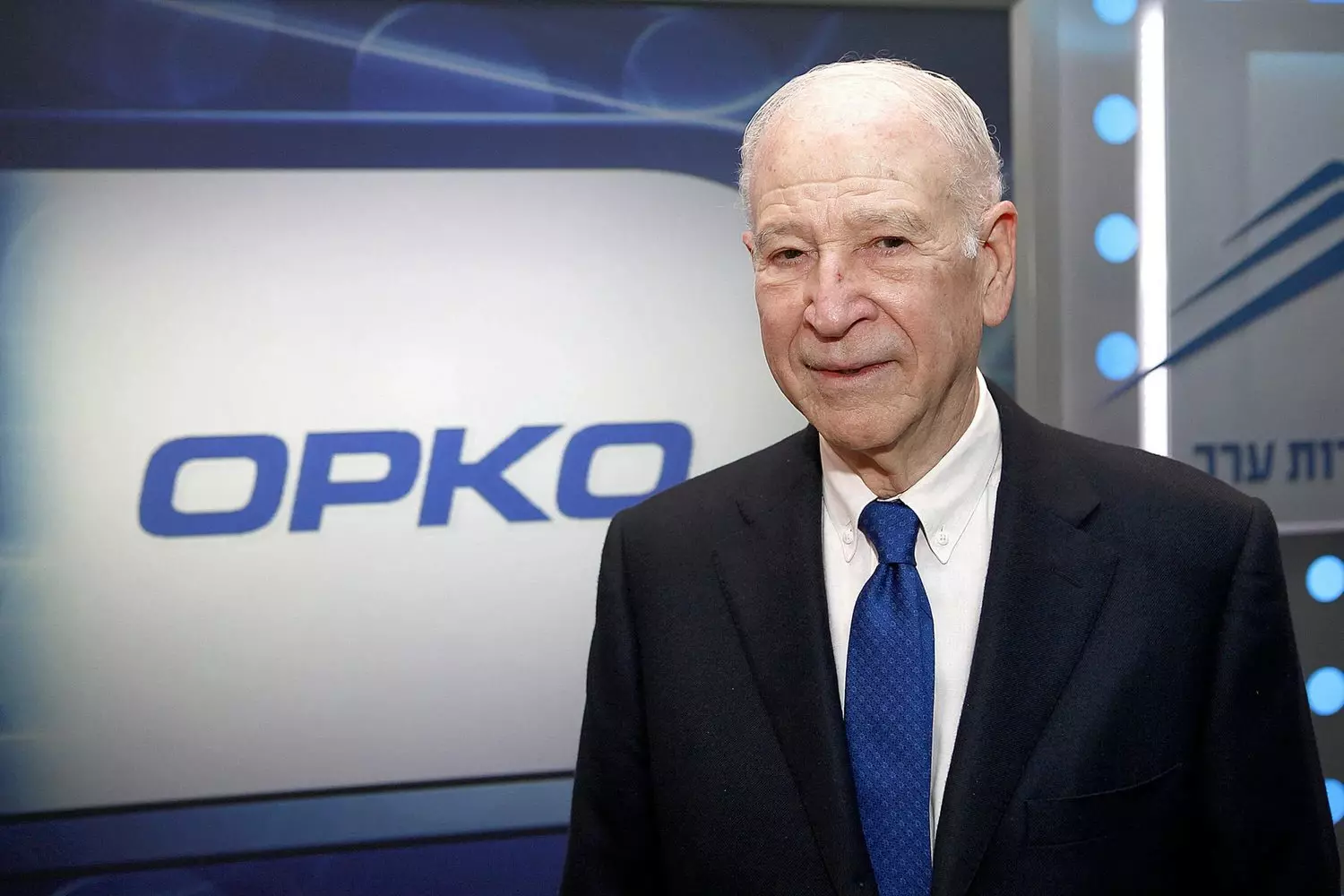 Phillip Frost – owner of OPKO Health, photo against a corporate background Phillip Frost in a suit with a blue tie, standing in front of the OPKO logo