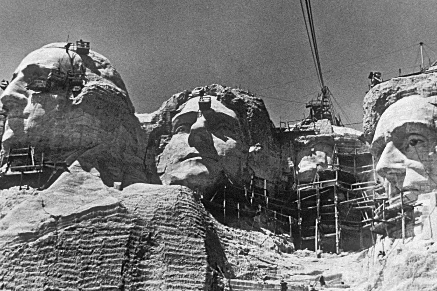 Under Construction: The Making of Mount Rushmore (1927-1941) Black and white historical photo showing workers carving the presidential faces into Mount Rushmore using ropes and pneumatic tools during the monument's construction era